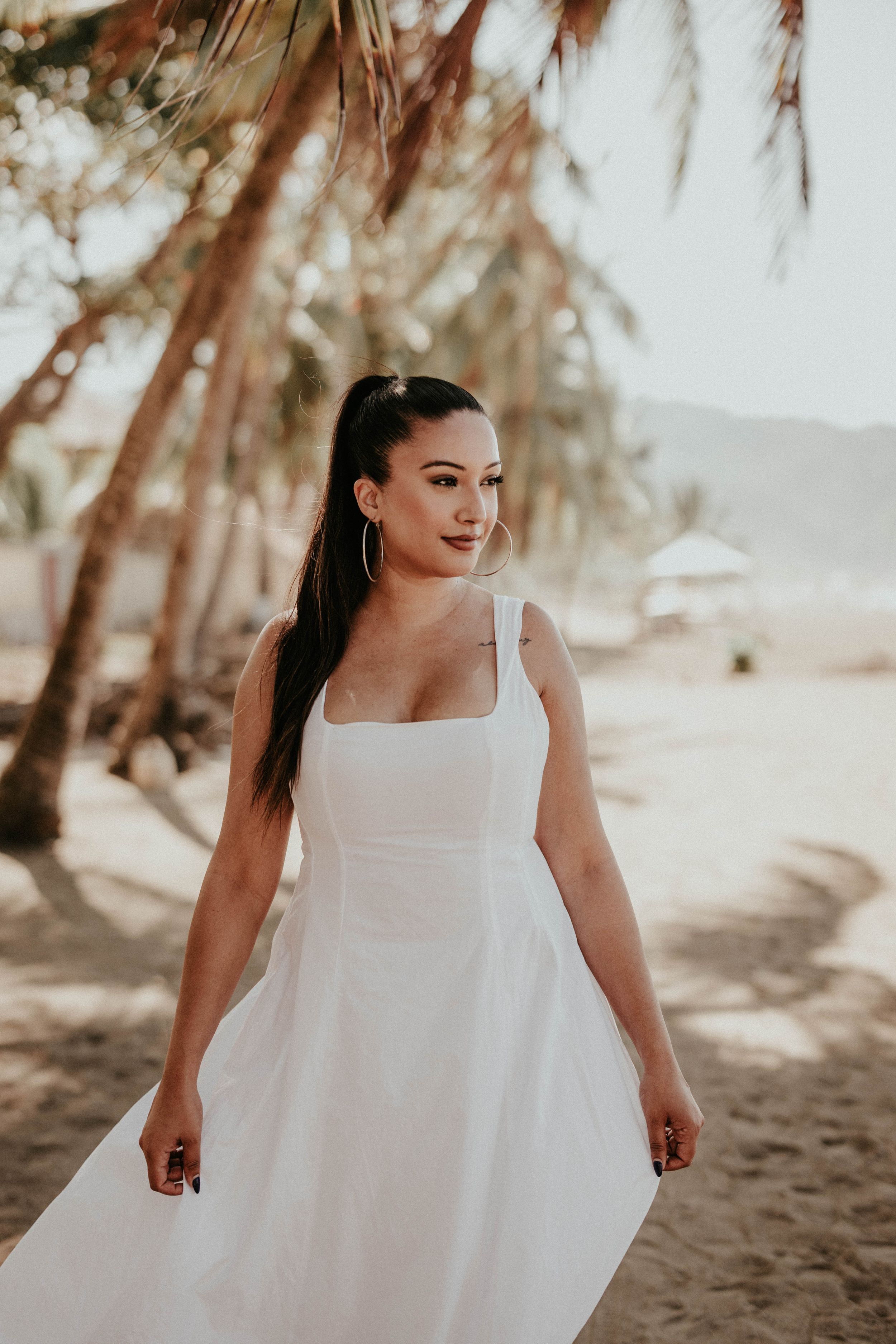 Woman in flowing white dress poses playfully on a tropical beach with palm trees in soft sunlight.