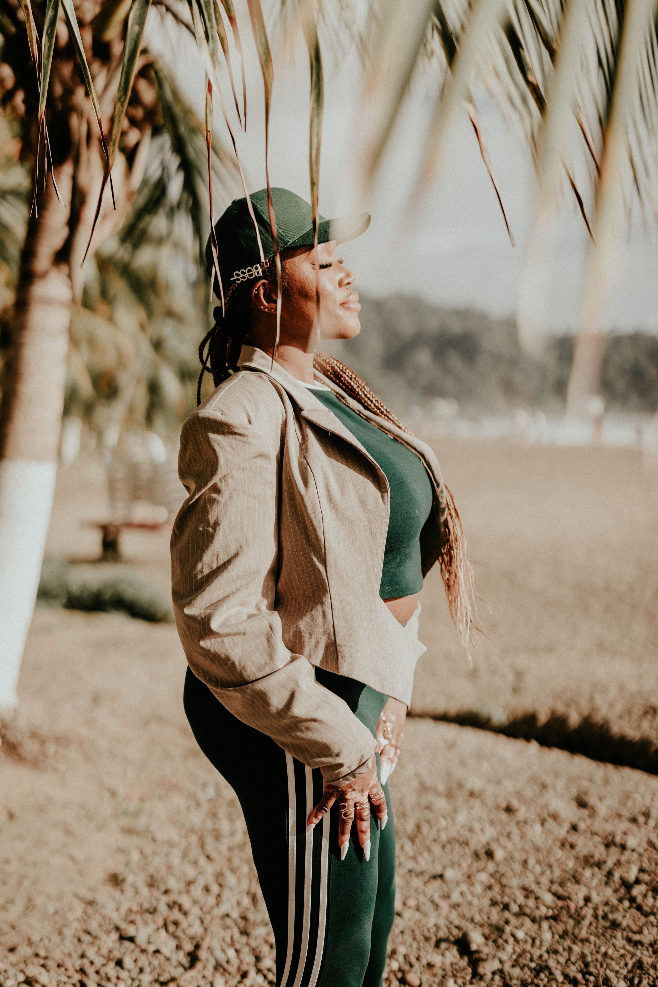 Person wearing a beige jacket and green pants poses thoughtfully among palm trees in warm sunlight.