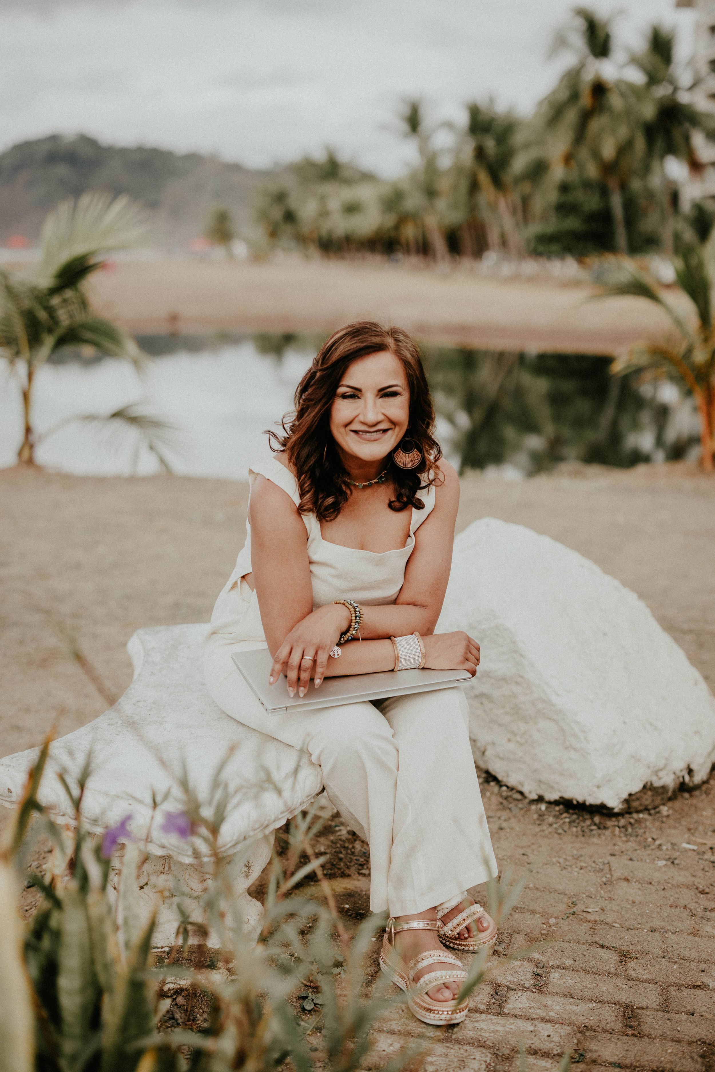 A person in a white wedding dress lies on a sandy beach surrounded by tropical palm trees and ocean views.