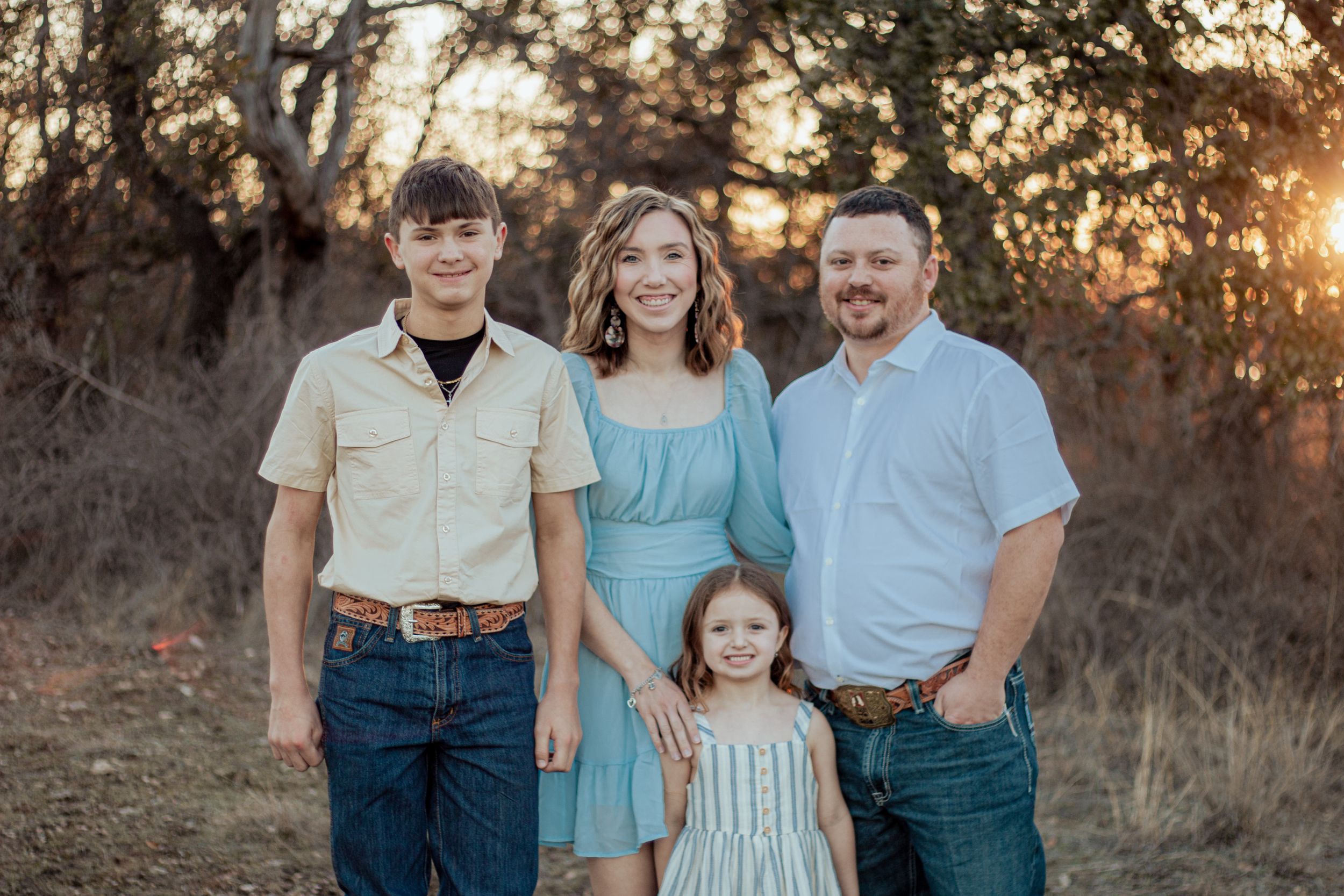 A family group in coordinating blue and white outfits poses together in a golden field at sunset.