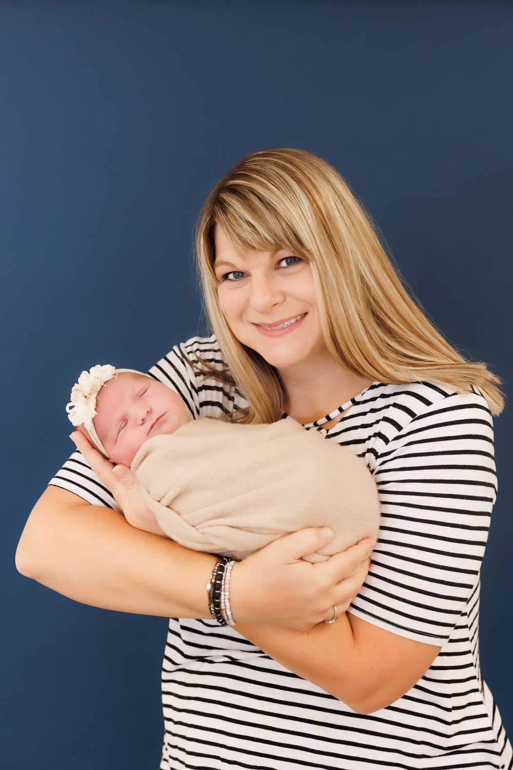 A woman in a striped shirt cradles a sleeping newborn baby against a dark blue background.