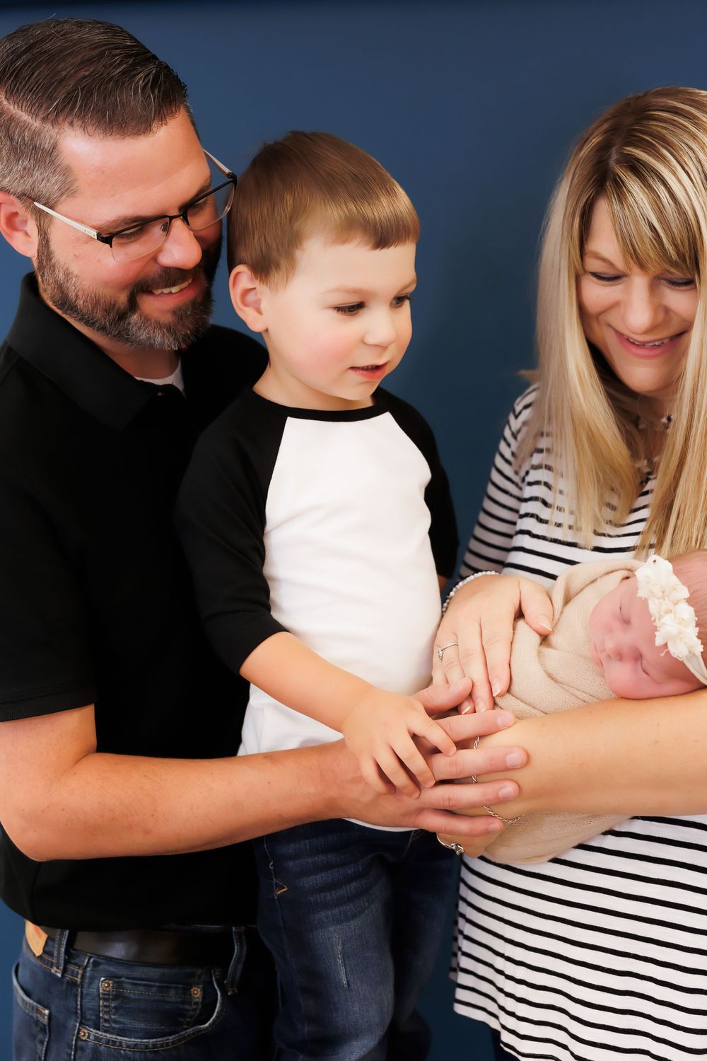 A family stands together admiring their newborn baby against a dark blue wall.