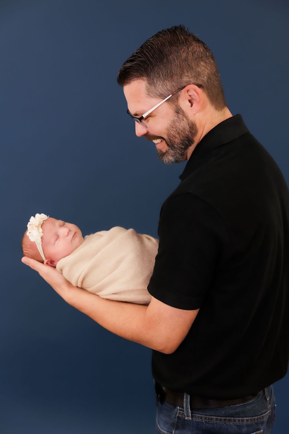A person in black shirt and jeans holds a newborn baby against a dark blue studio backdrop.