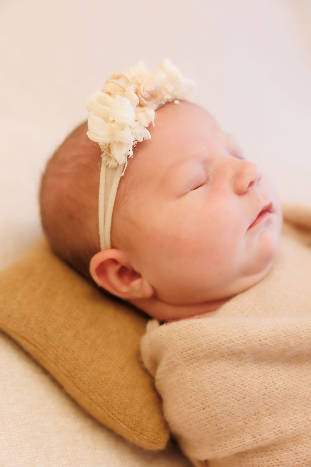 Sleeping baby wearing cream colored outfit and delicate floral headband rests on tan pillow.