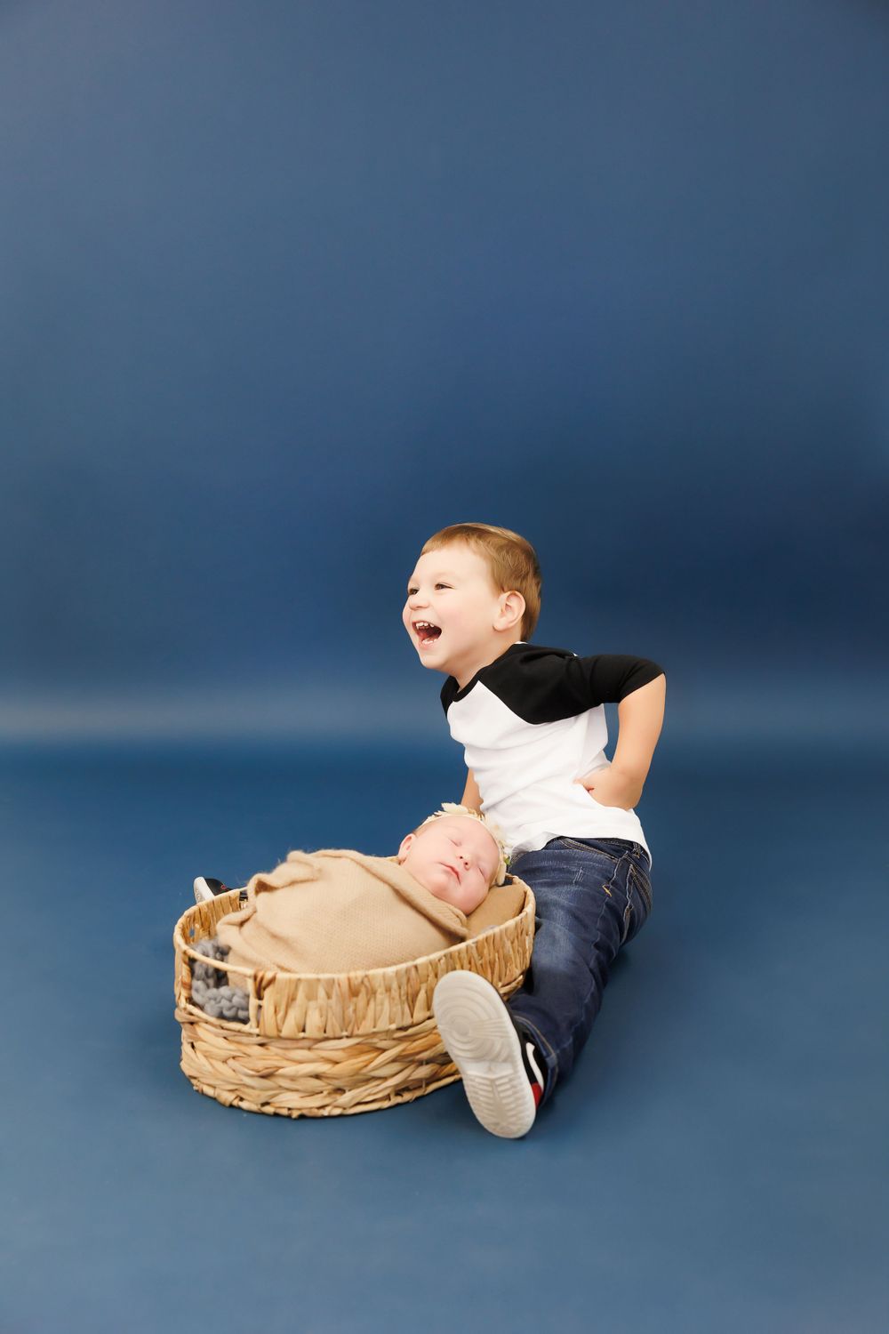 Young child laughing while sitting next to a wicker basket against a blue studio backdrop.