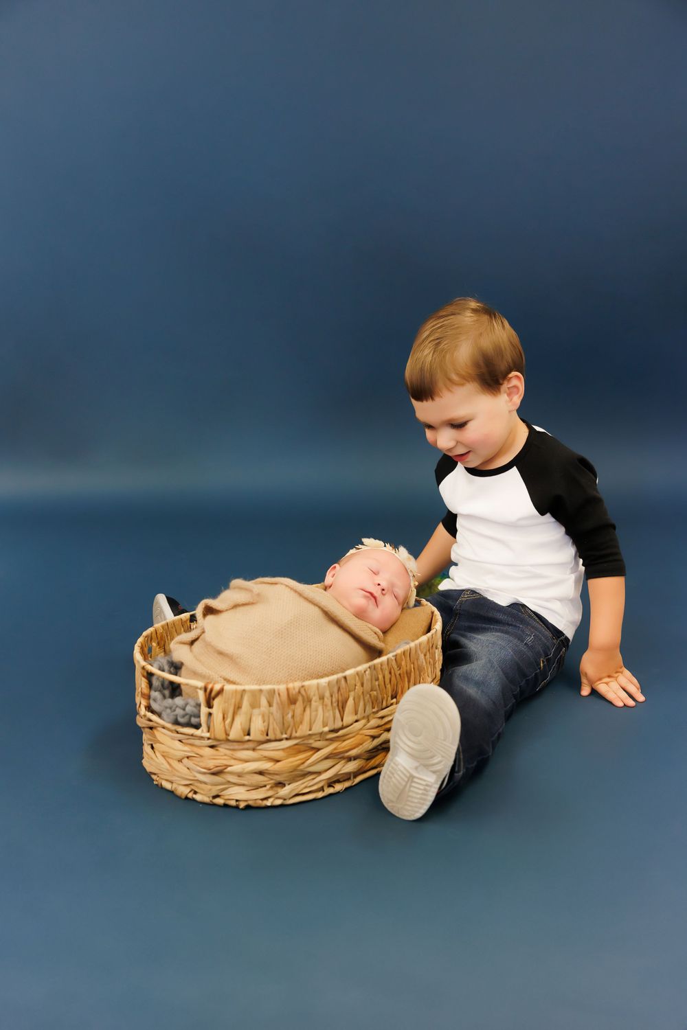 Young boy in black and white shirt sits next to baby in wicker basket against blue studio backdrop.