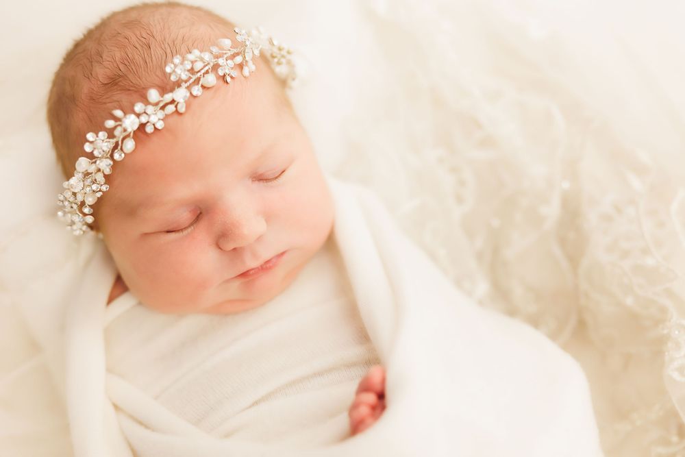 Sleeping baby wrapped in white swaddle wearing delicate pearl and crystal headband on soft cream background.