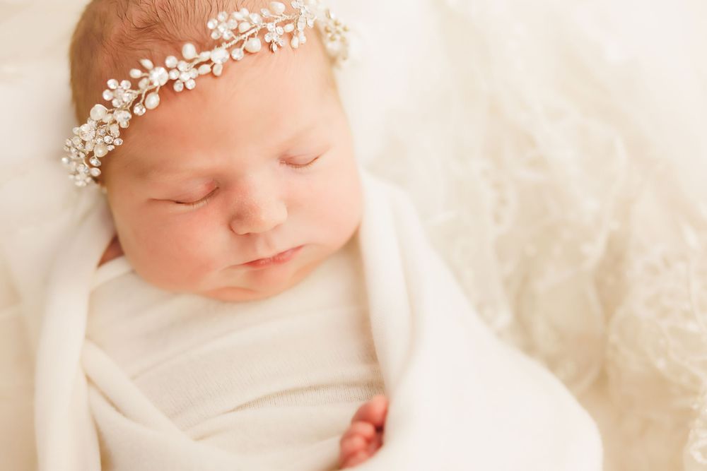 Peaceful sleeping newborn wearing delicate floral headband wrapped in soft white blanket.