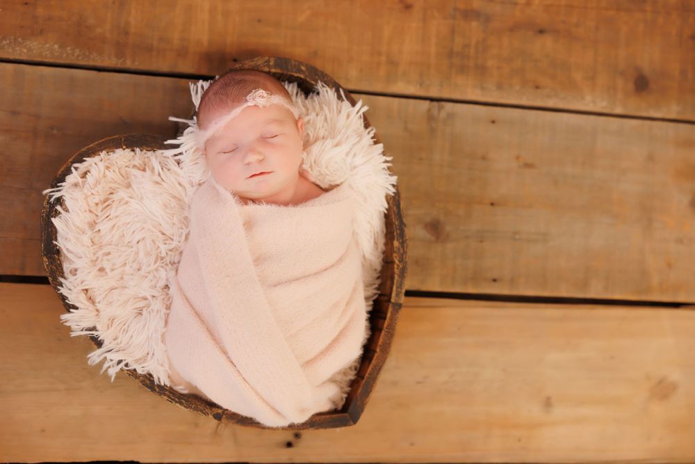 A sleeping newborn wrapped in pink swaddle blanket lies in heart-shaped fur nest on wooden background.