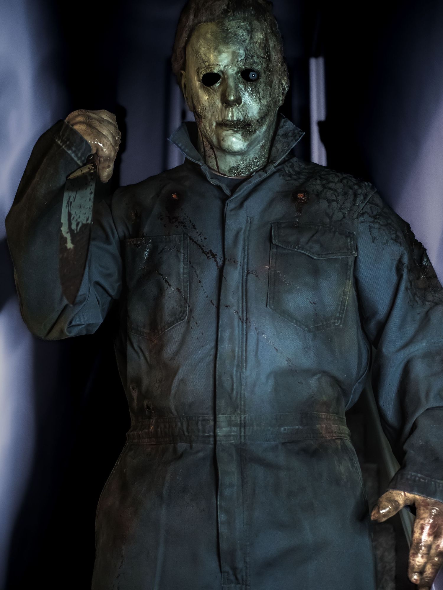 A menacing figure in dark coveralls and a weathered hockey mask stands in dim lighting holding a threatening pose.