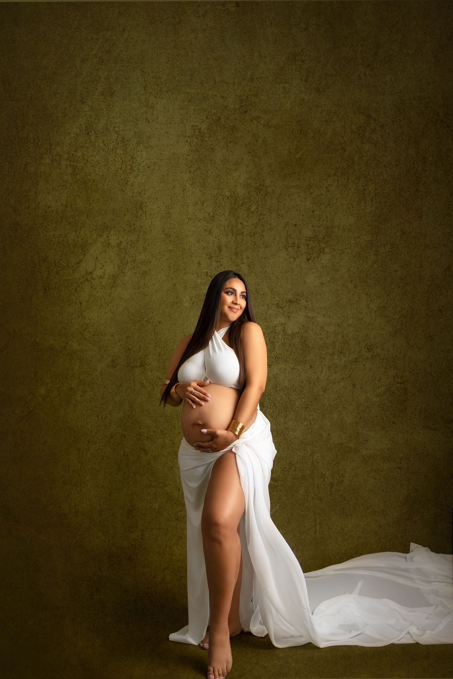 Artistic maternity portrait featuring an expectant mother in flowing white fabric against a textured olive green backdrop.