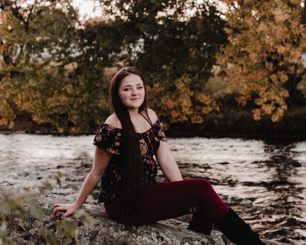 Outdoor senior photography session by water featuring dark floral dress and natural autumn scenery.