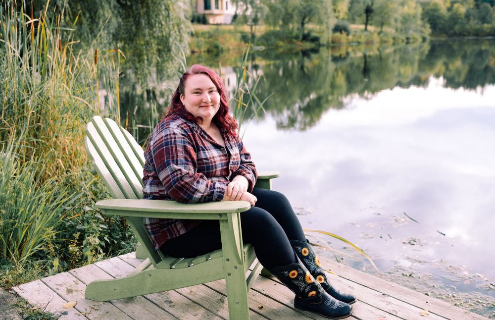 Person relaxing in an Adirondack chair on a wooden dock surrounded by autumn foliage.