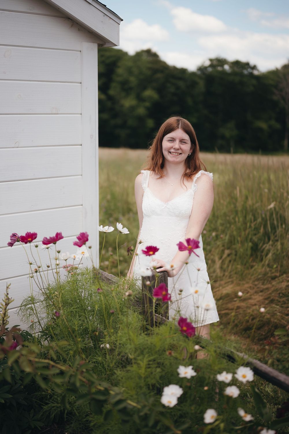 Young woman in white dress smiling among pink and white cosmos flowers beside a white shed in a green field.