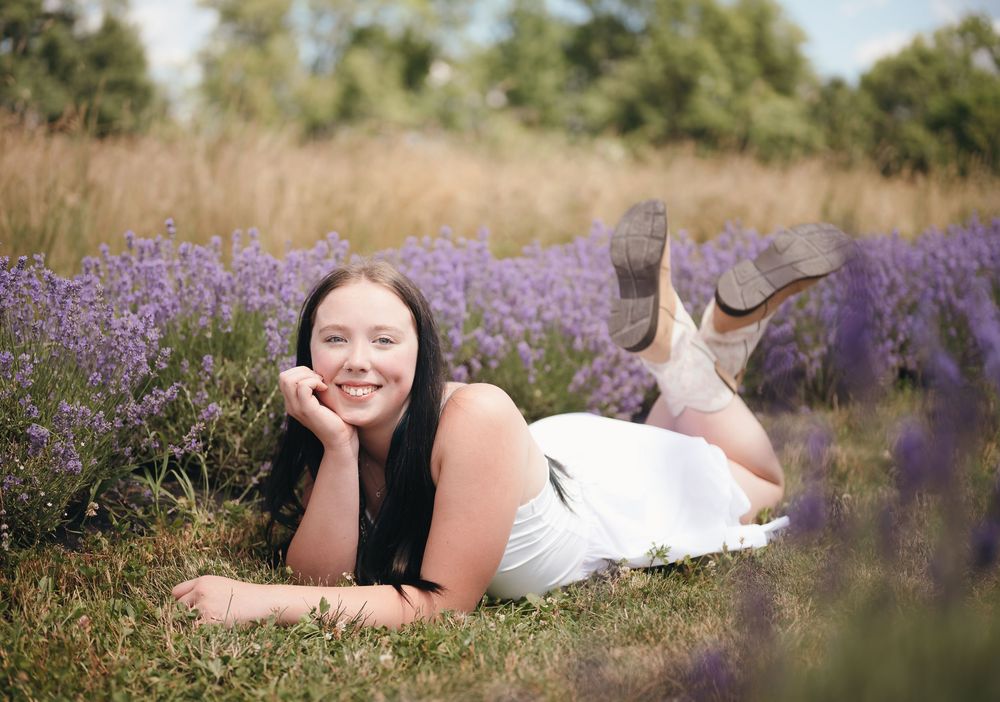 Young woman in white dress lying in purple lavender field, smiling with hand on chin, cowboy boots visible.