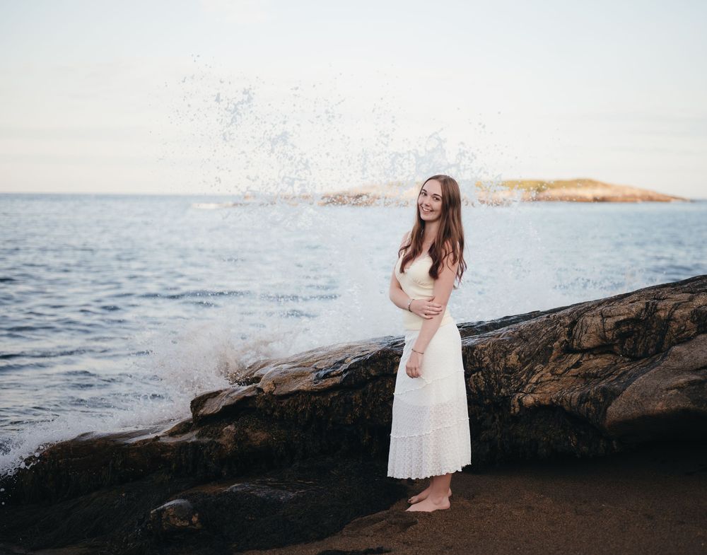 Young woman in white dress standing on rocky shore with ocean waves splashing behind her at sunset.