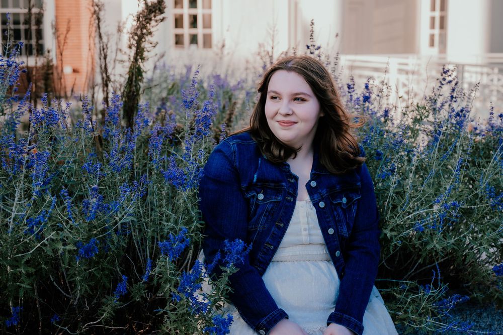 A person in a navy blue coat poses among vibrant purple wildflowers in a garden setting.