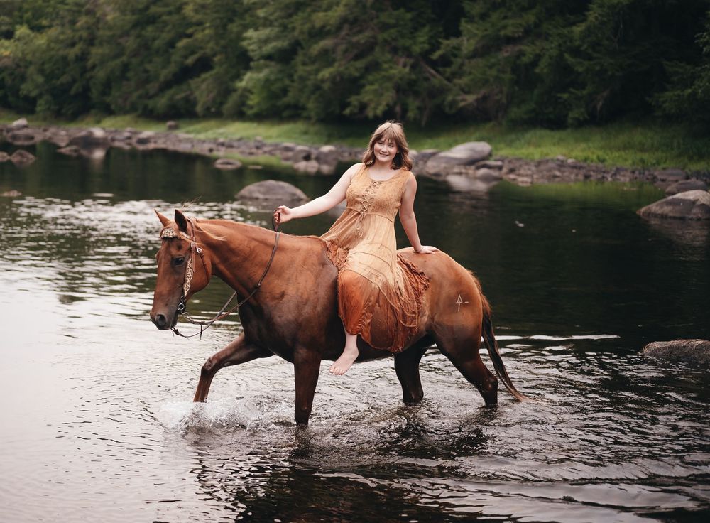 Woman in golden dress riding a chestnut horse through a shallow river with lush green trees in background.
