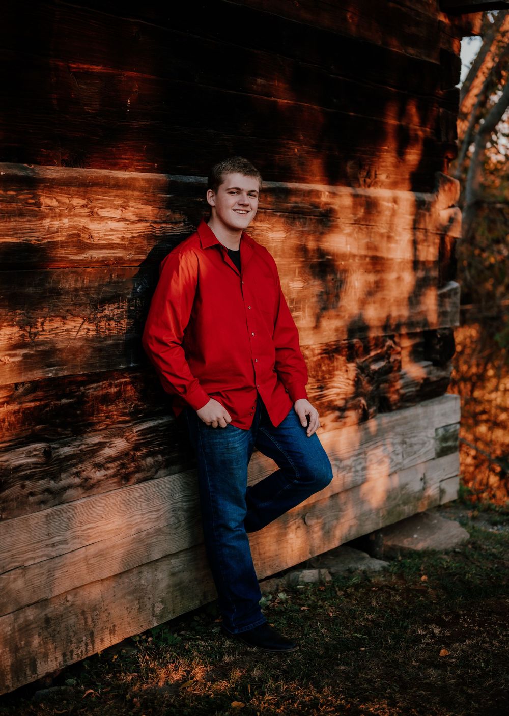 Person in a red shirt and jeans sits against a rustic wooden wall in warm sunset lighting.