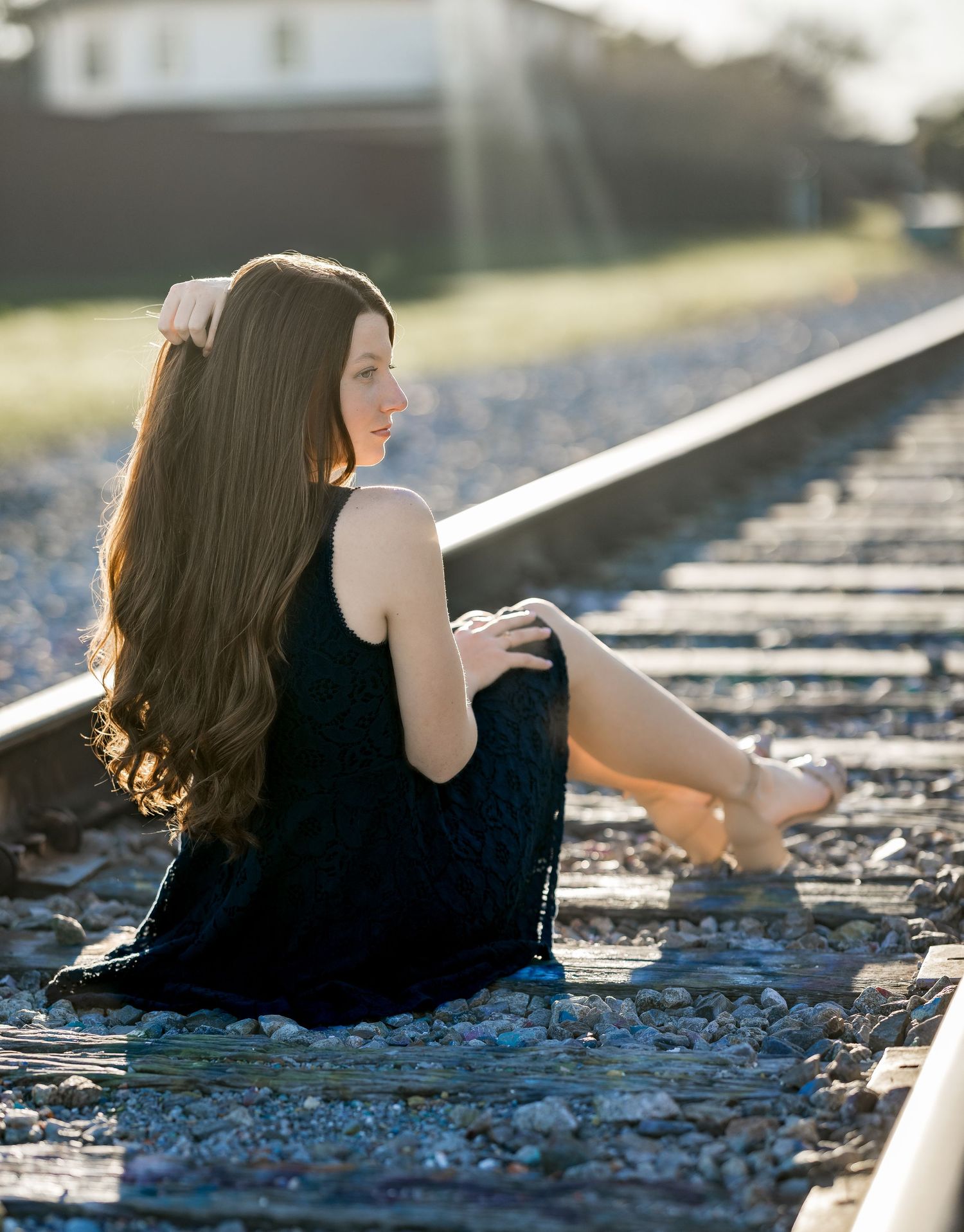 A figure in a black dress sits on railroad tracks during golden hour, with long dark hair flowing in the sunlight.