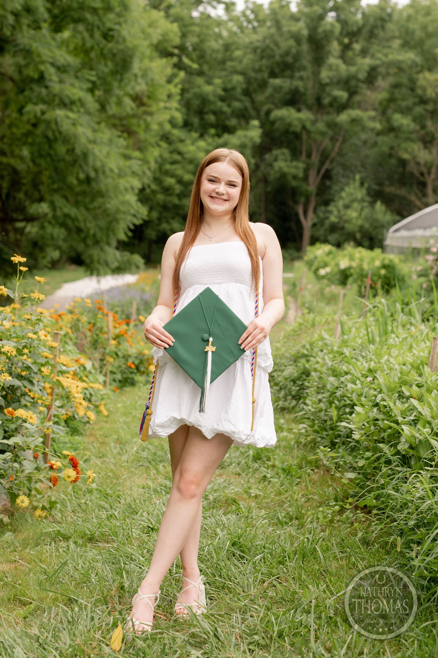 Graduate holding green diploma folder poses in white dress surrounded by wildflowers.