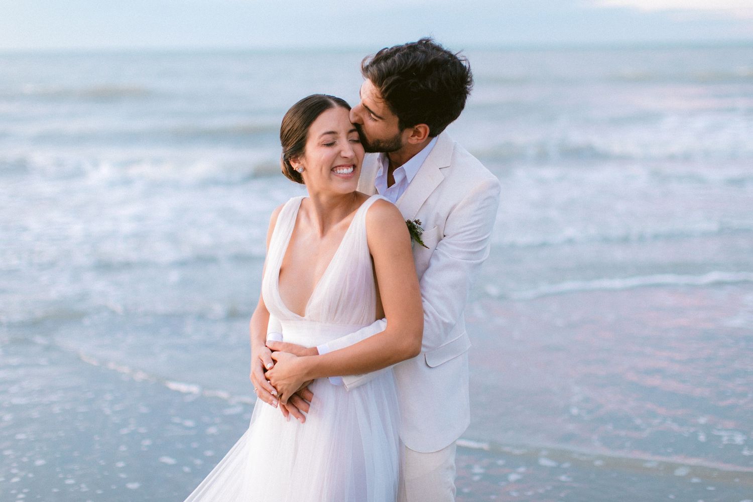 A romantic couple embraces on the beach at sunset, with the bride wearing a flowing white dress.