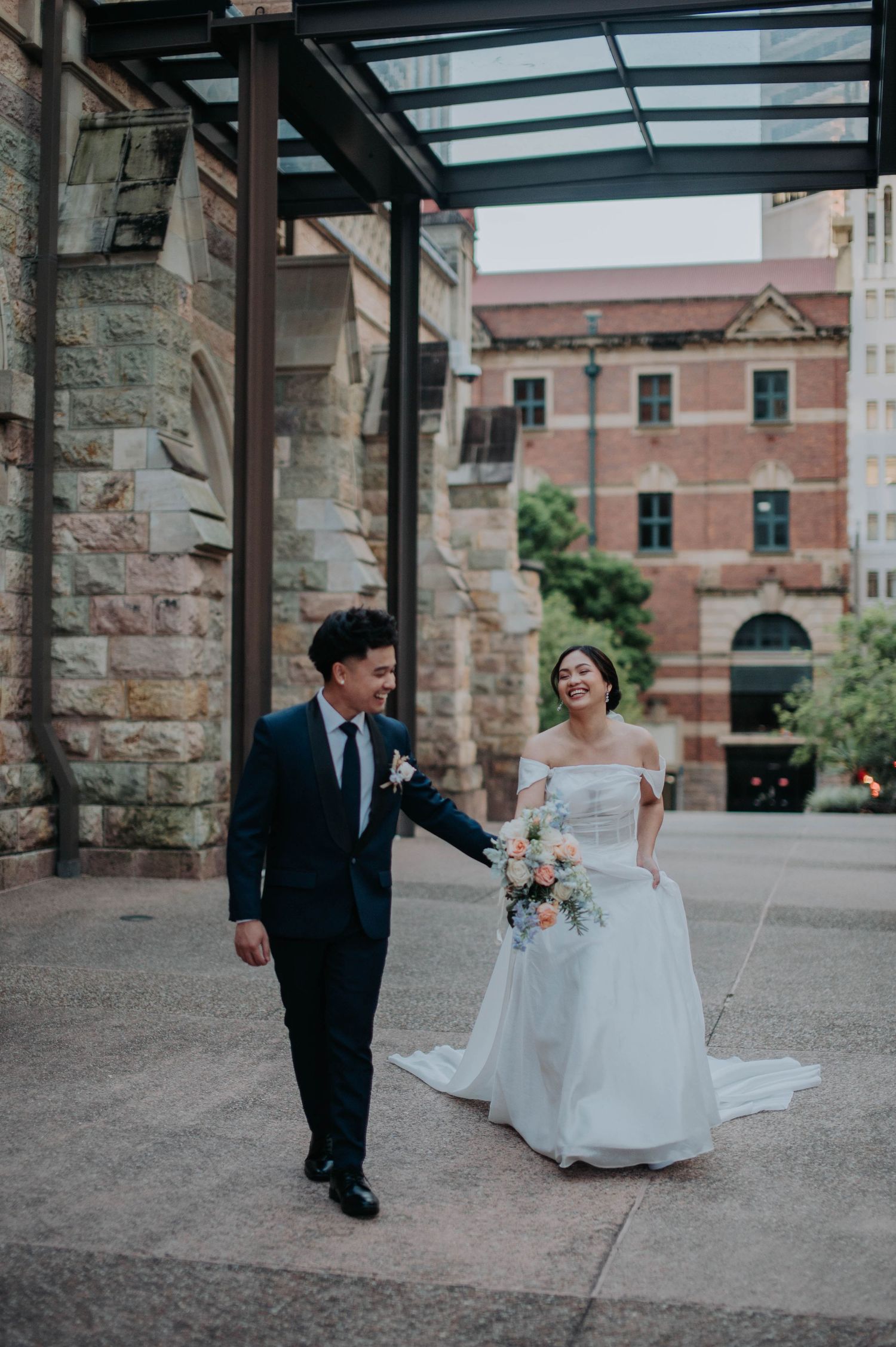 A couple strolls under a glass walkway canopy beside a historic stone building.