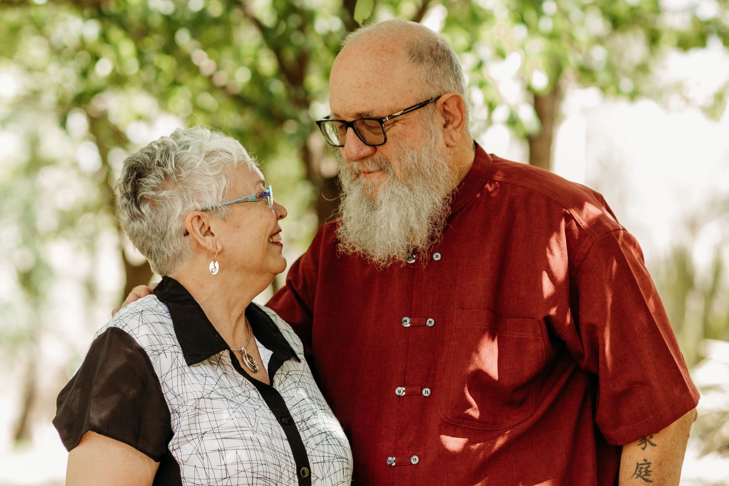 An elderly couple sharing a tender moment outdoors, with the man in a red shirt and the woman in a black and white top.