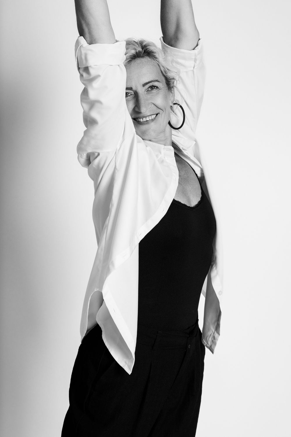 Black and white photo of smiling woman in white shirt and black outfit, arms raised overhead in studio setting.