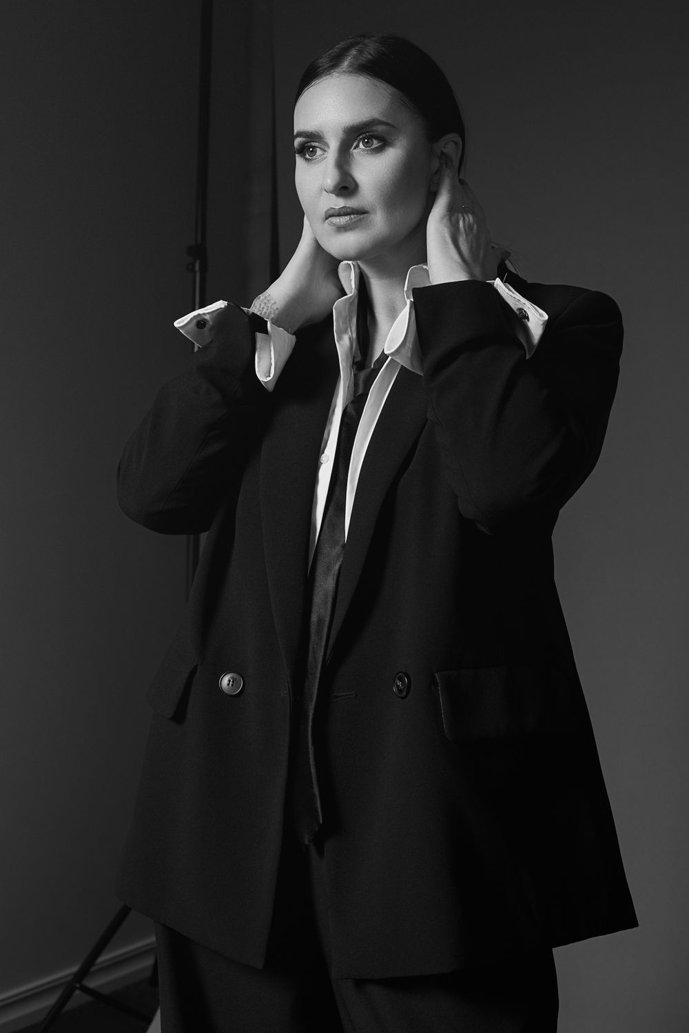 Black and white portrait of an elegant woman in a blazer and dress shirt, hands raised to her neck, posing confidently.