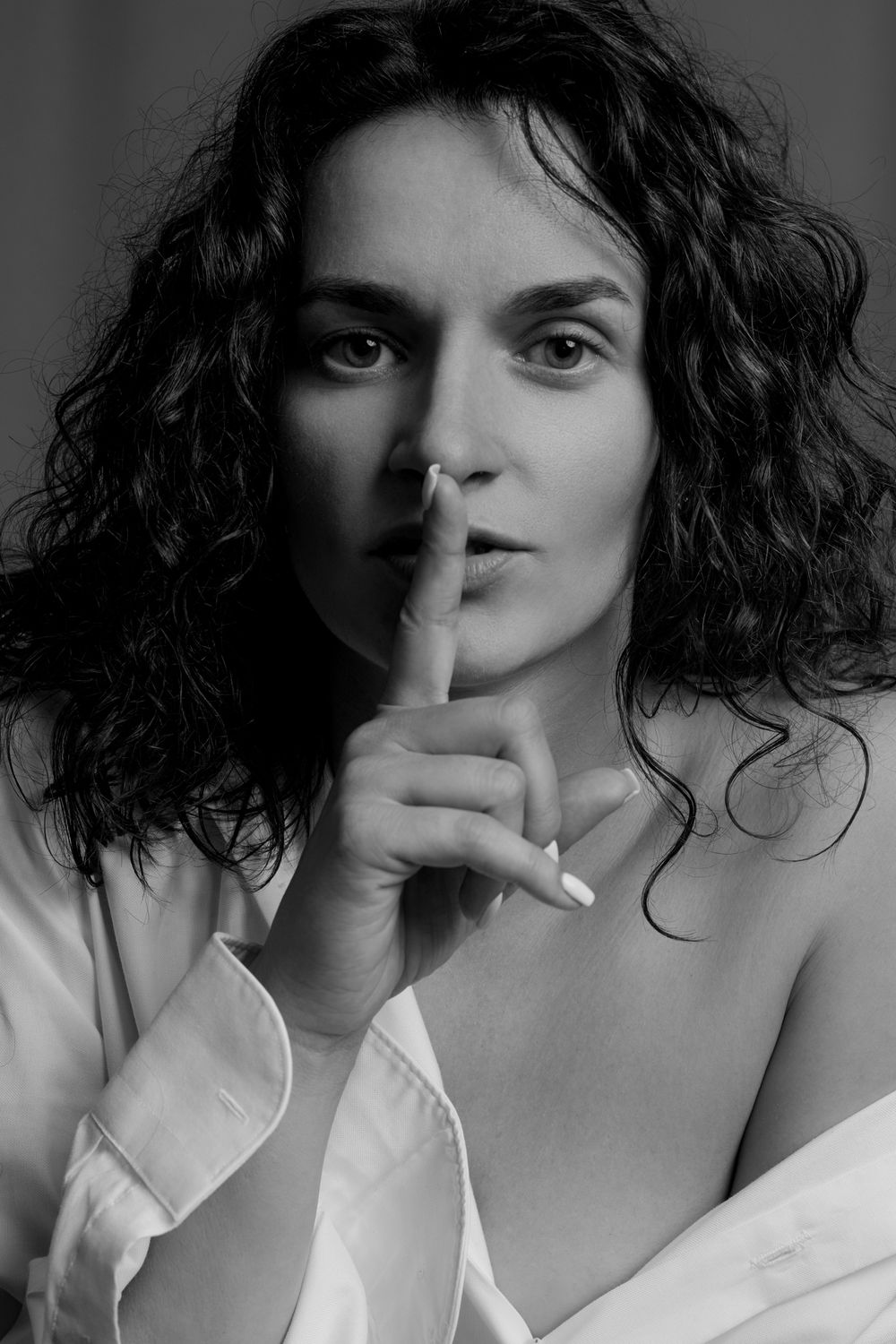 Black and white portrait of a woman with curly hair making a shush gesture with finger to lips.