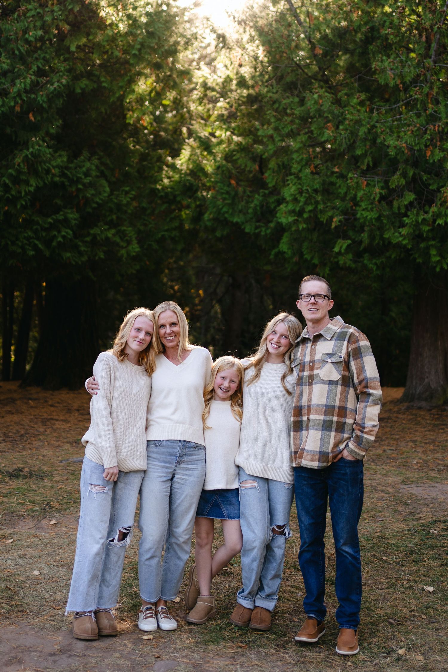 Family wearing coordinated casual outfits stands together on a dirt path with green trees and golden sunlight behind them.
