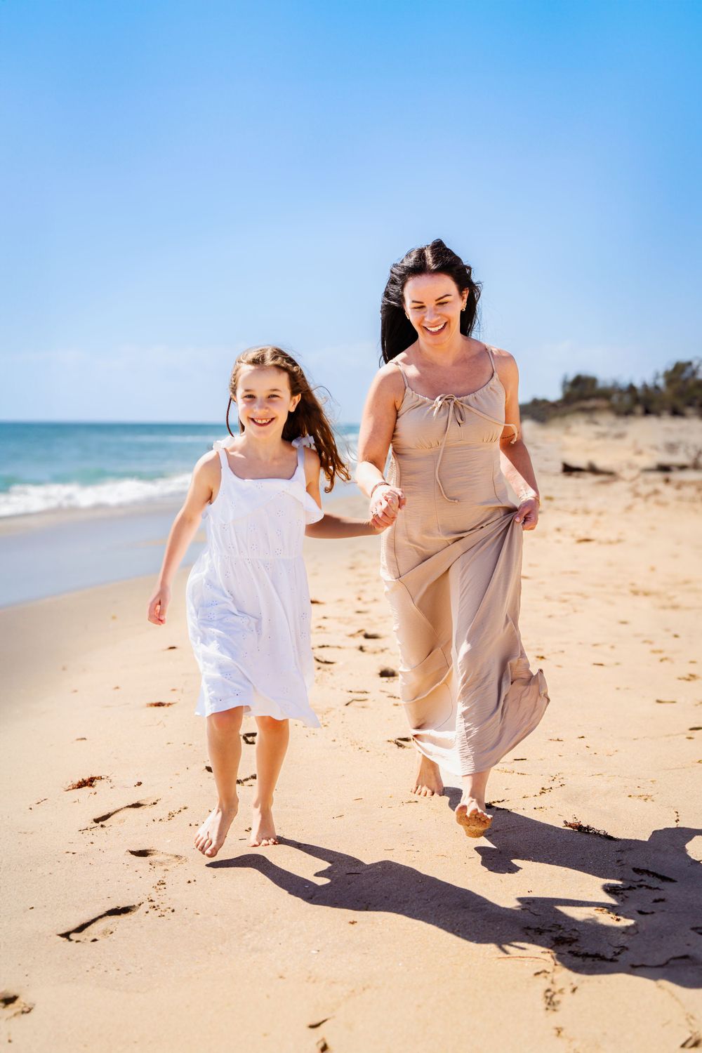 Woman and child holding hands while running joyfully on beach with waves in background.
