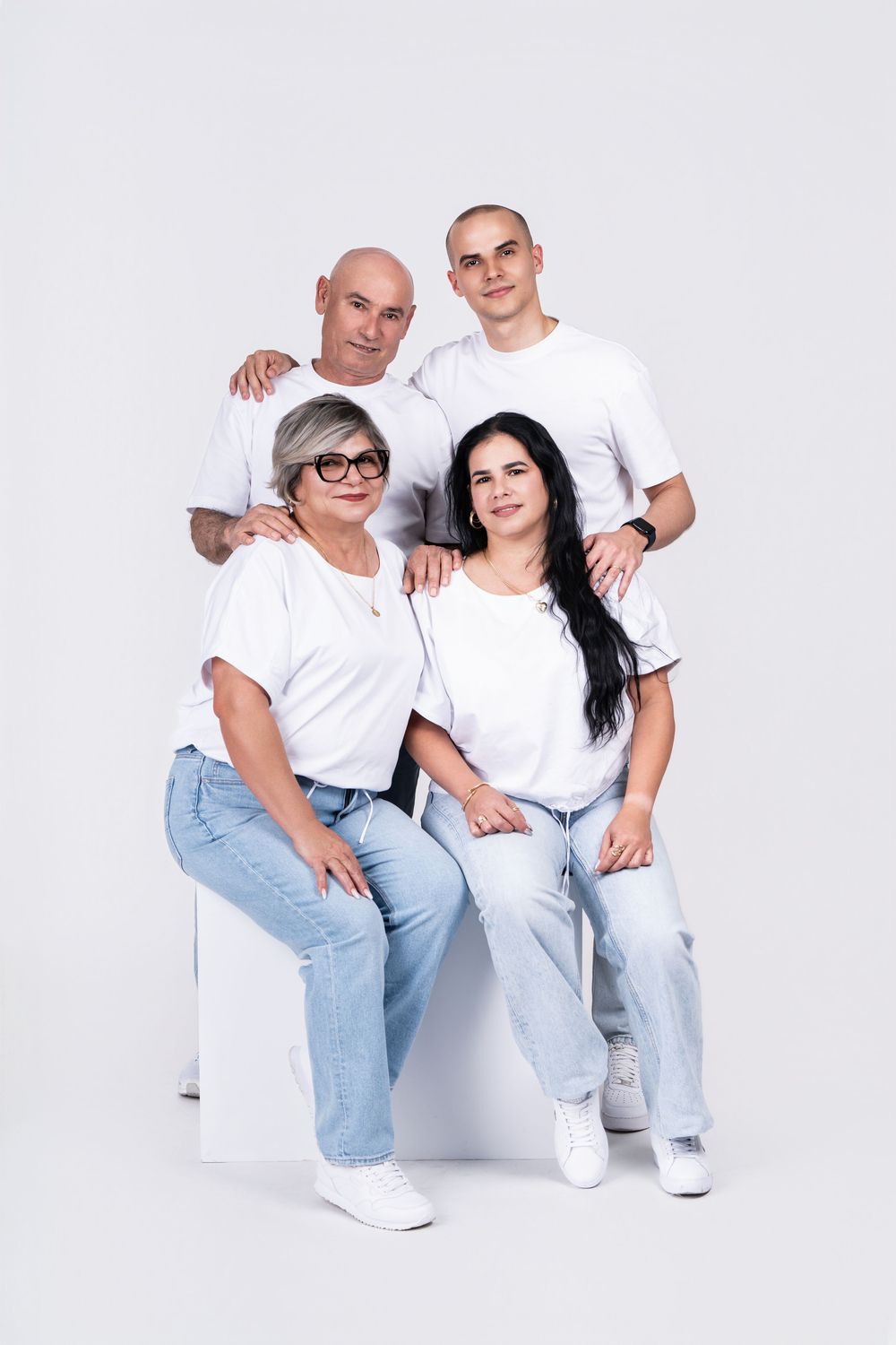 Family group wearing coordinated white tops and jeans pose casually in a studio setting.