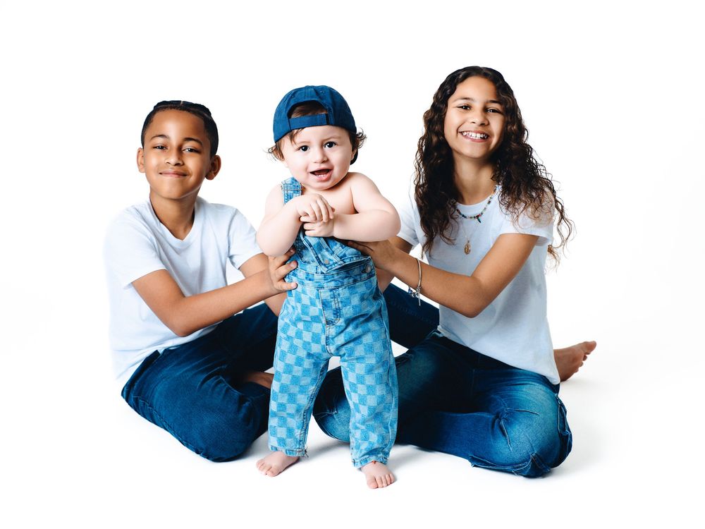 Two older siblings sit on white background smiling while holding a happy baby in blue checkered outfit and cap.