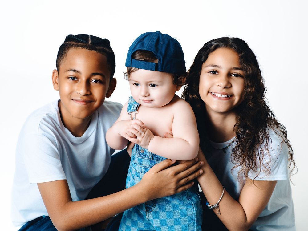 Two older children smile while holding a baby wearing a blue checkered outfit and cap against a white background.