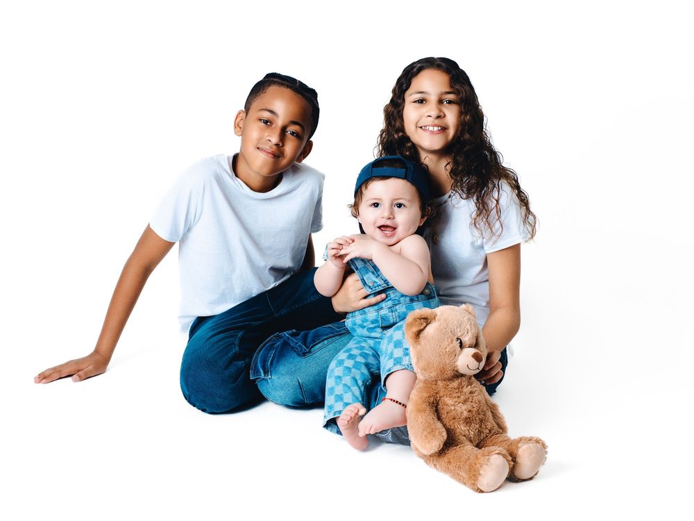 Two siblings and a baby pose with a teddy bear on a white background in a cheerful family portrait.