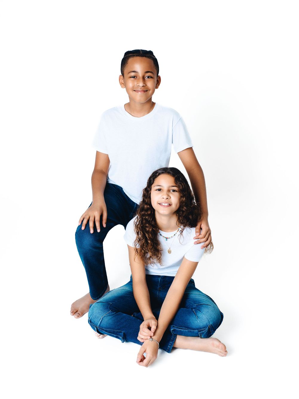 Two smiling children in white t-shirts and jeans pose together against a white background in a studio portrait.