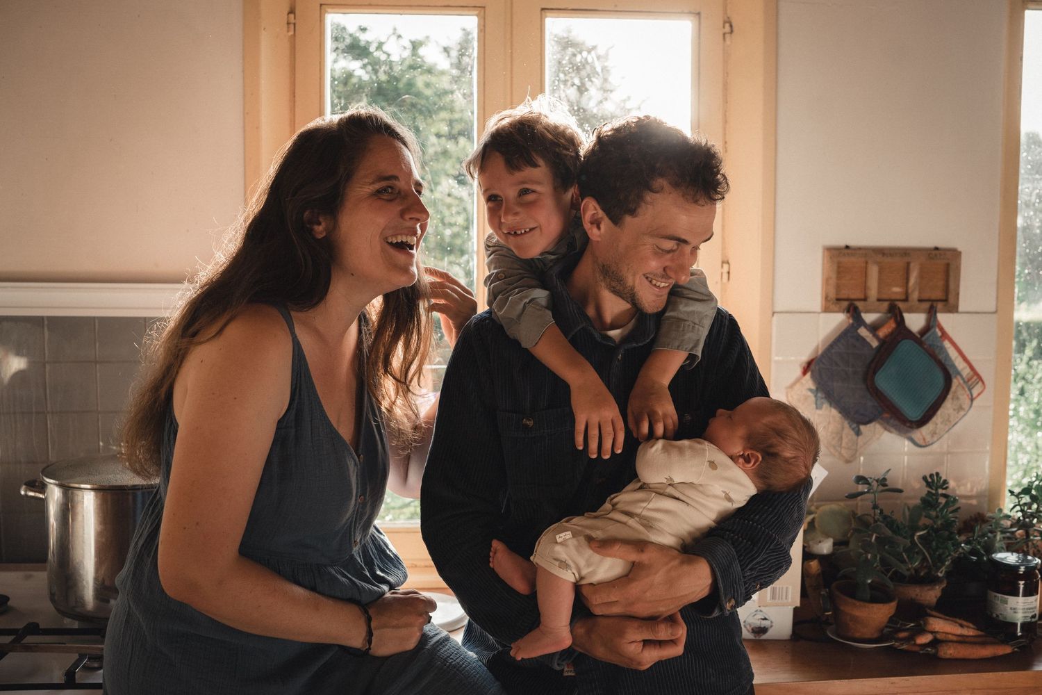 Seance photo famille dans une cuisine maison de campagne couple parent souriant avec leur fils debout enlaçant le père qui tient son bébé