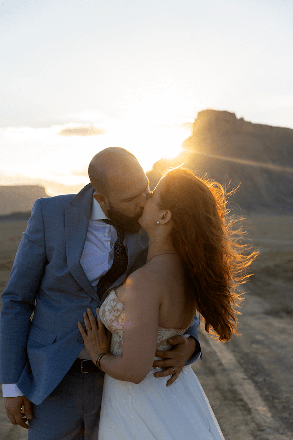A romantic couple shares an intimate kiss at sunset in the hills while wearing wedding attire.
