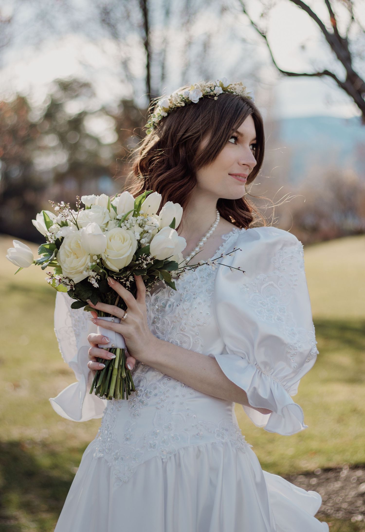 Bride in white lace dress holding white rose bouquet in outdoor garden setting.