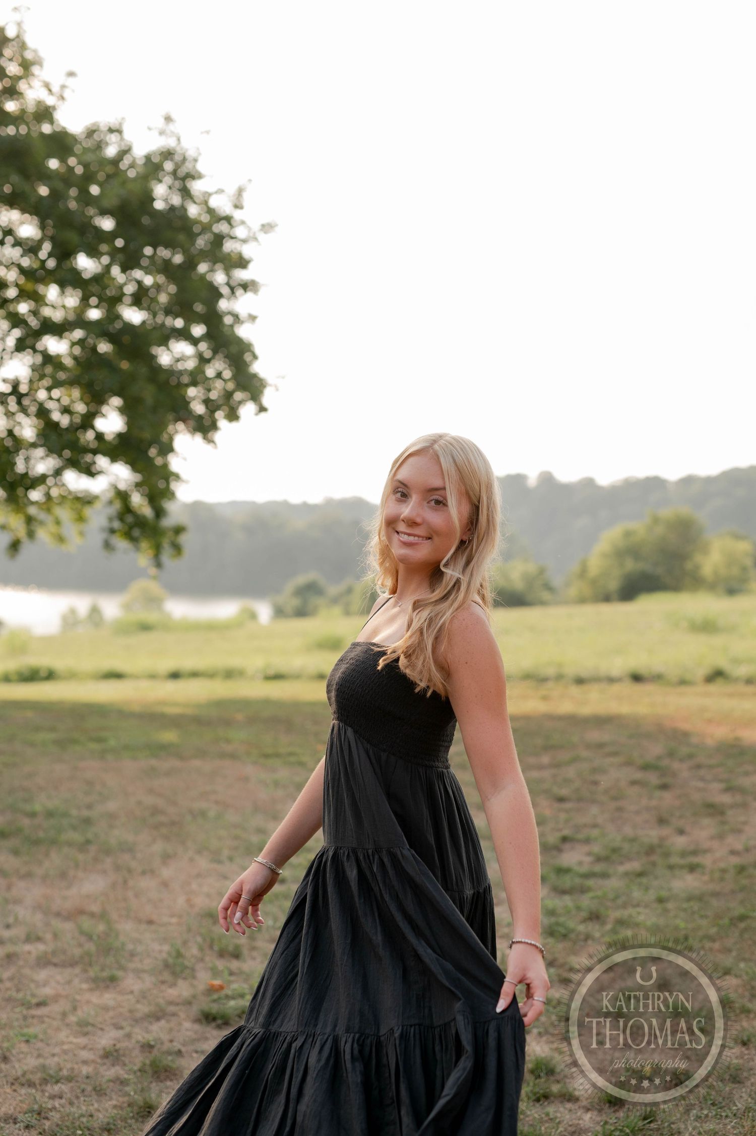 A woman wearing a black maxi dress poses in a sunlit meadow with trees and rolling hills in the background.
