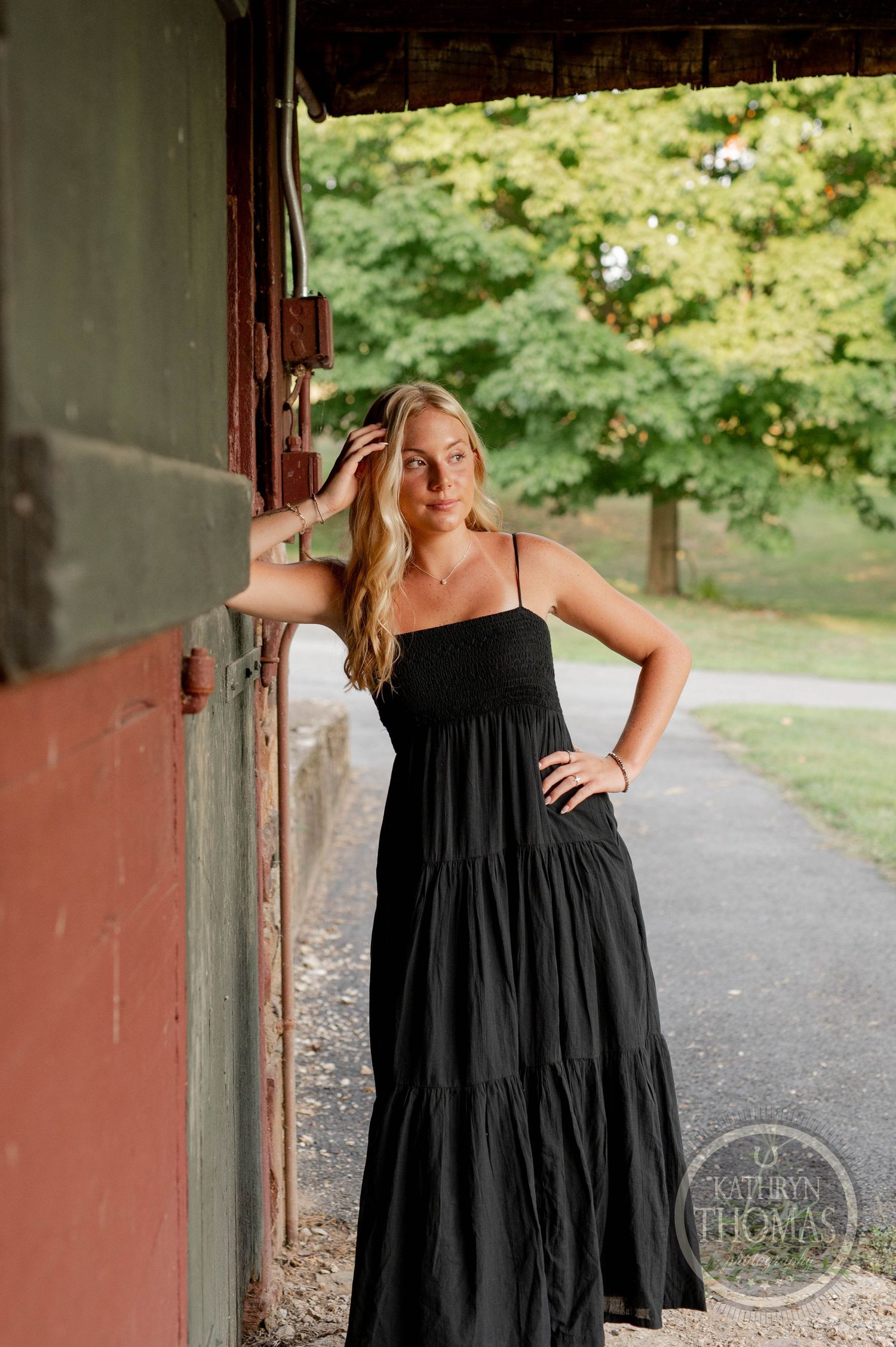 A woman in a tiered black maxi dress poses against a red barn wall during a summer photoshoot.