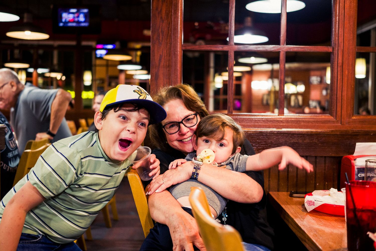Three people pose playfully while dining at a casual restaurant with dark wood paneling and overhead lighting.