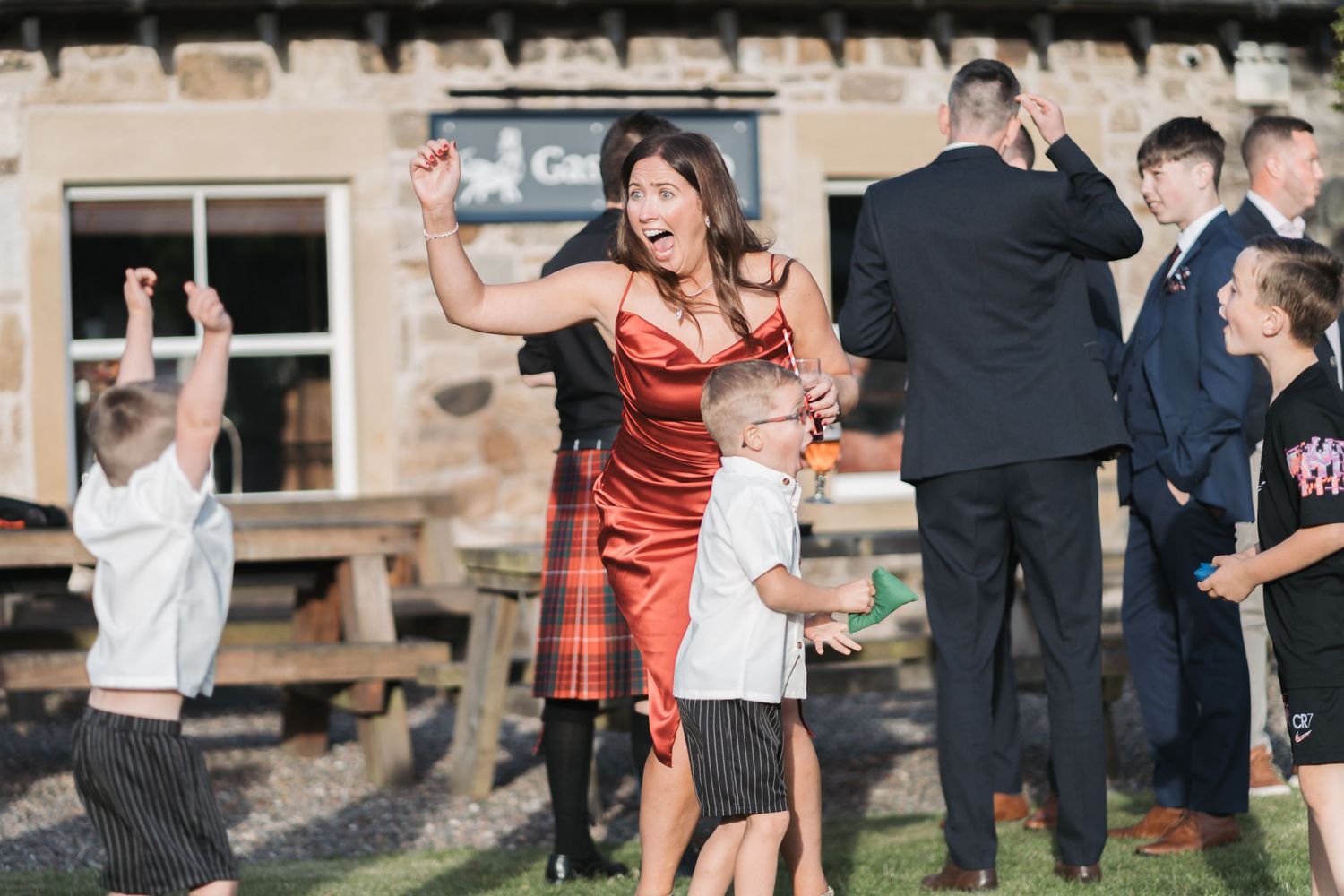 People dancing and celebrating at an outdoor wedding reception venue with rustic wooden buildings in the background.