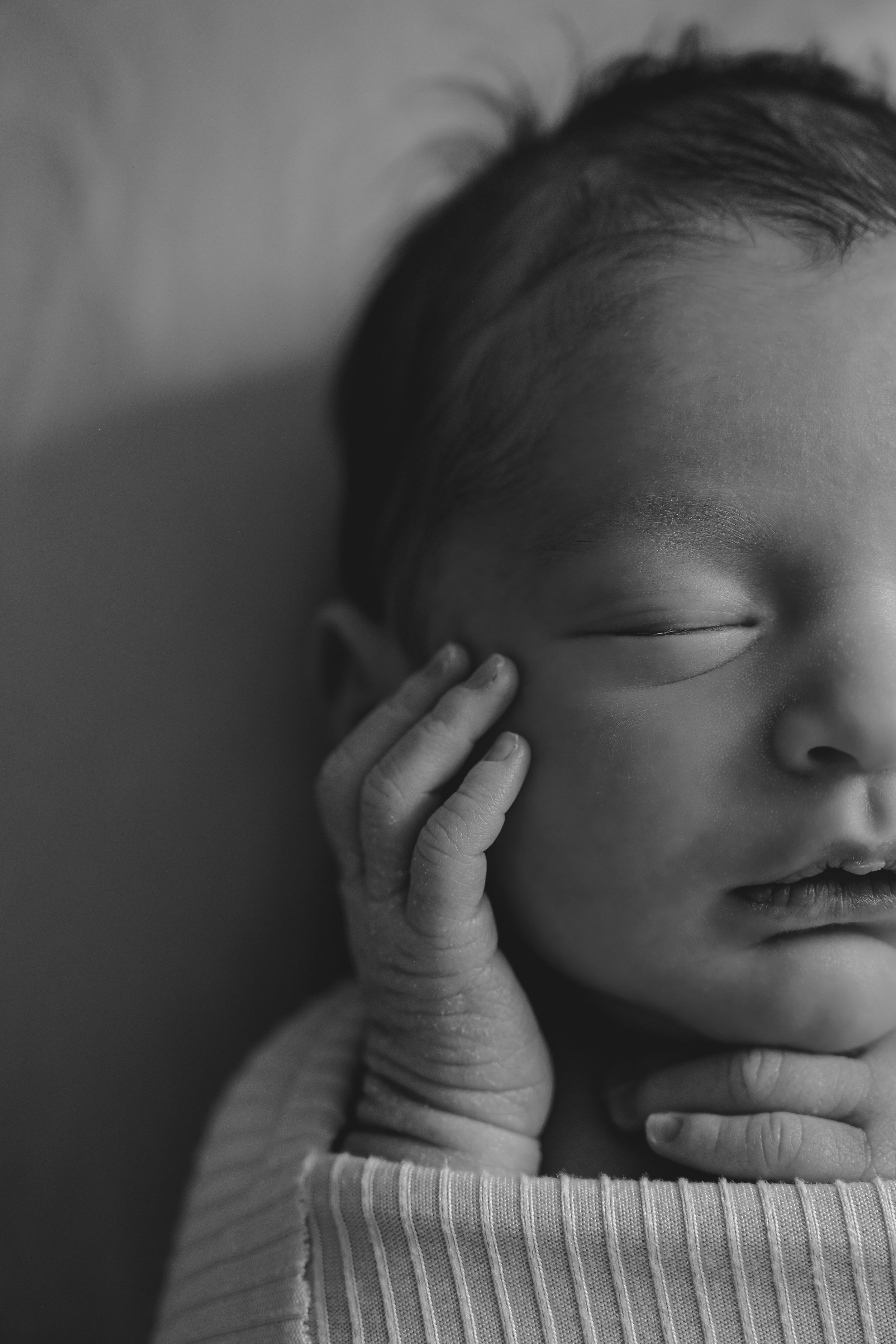 Black and white close-up photo of a sleeping newborn baby with tiny hand raised near face.