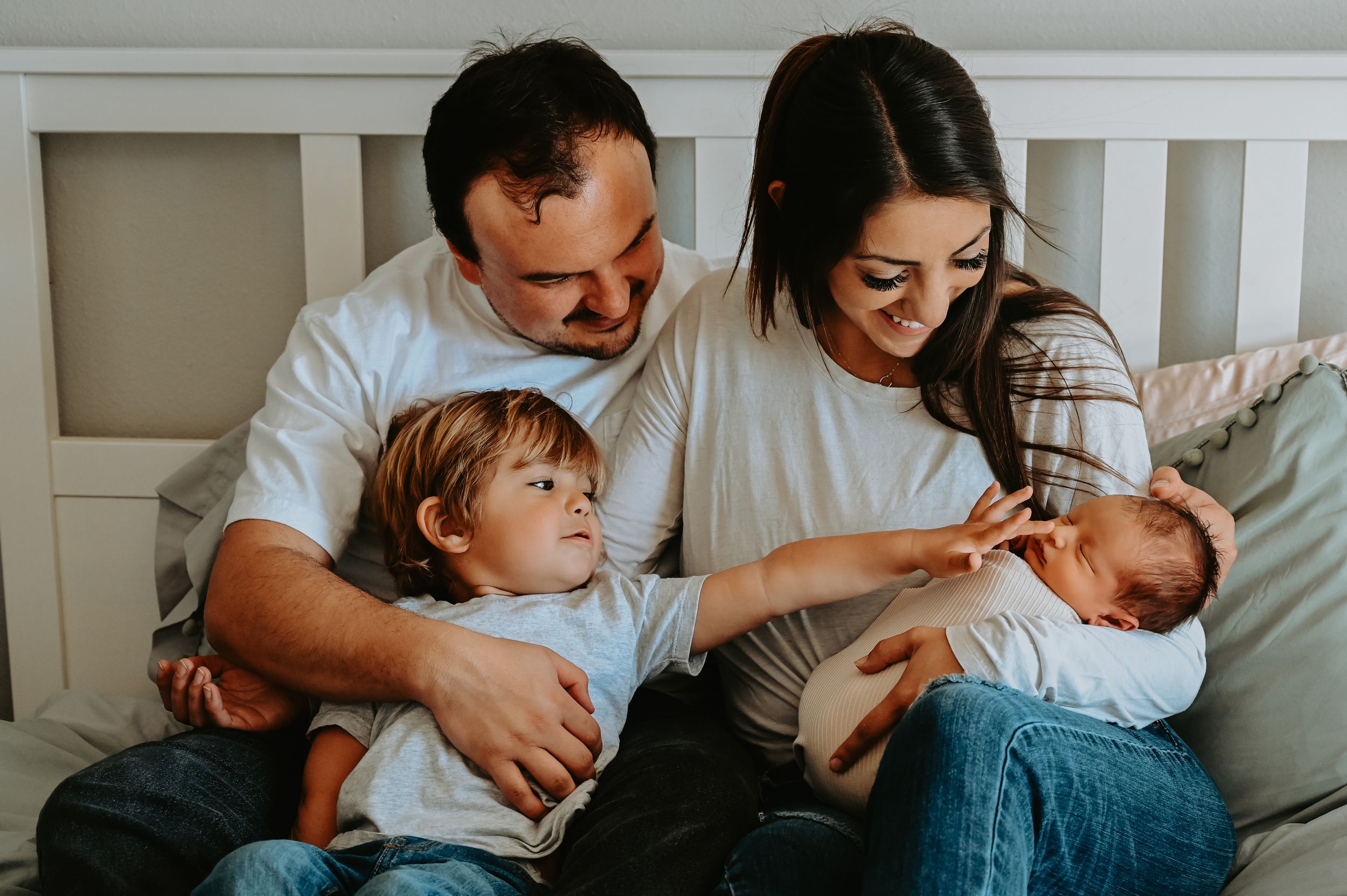 Family of four sitting on bed, parents smiling at newborn baby while toddler looks on curiously.