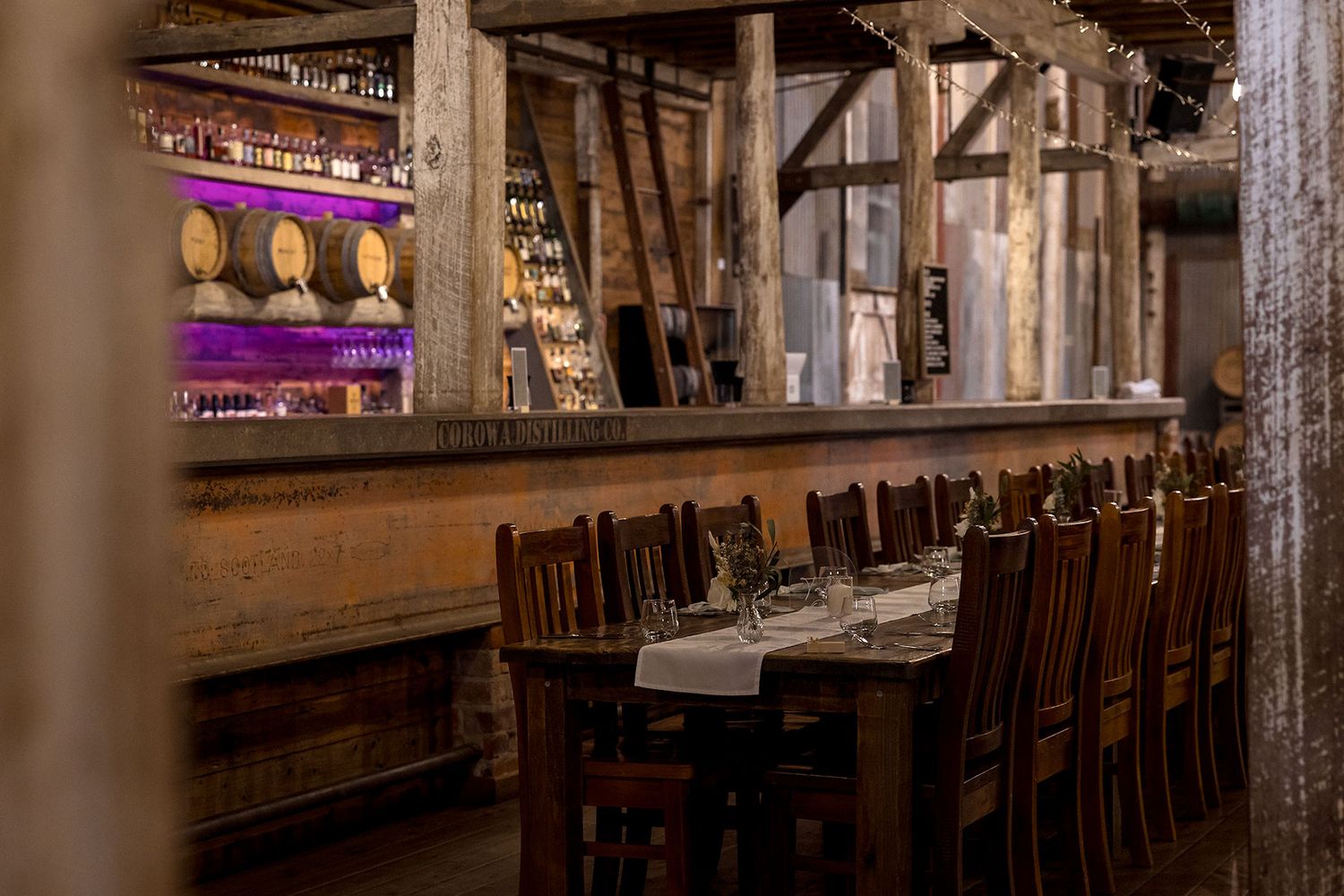 Long rustic dining table with wooden chairs set in an industrial warehouse space with exposed beams and warm lighting at Corowa Distilling.