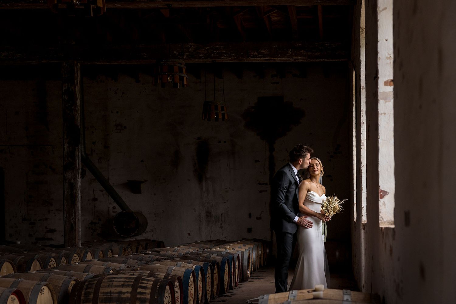 A couple stands in romantic embrace between rustic wooden barrels in a dimly lit winery barrel room at the Corowa Distilling Company.