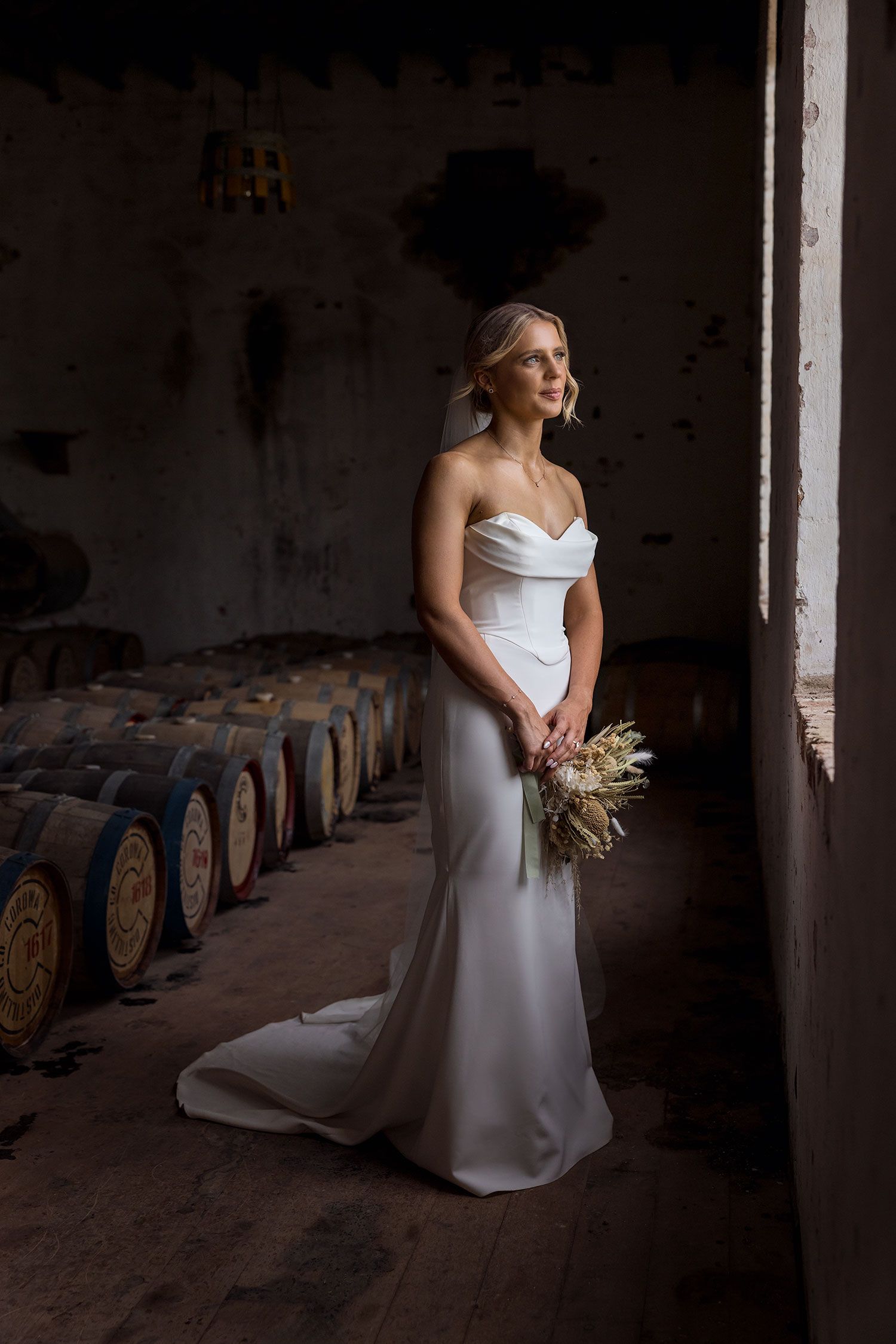 Bride in elegant white strapless gown from LUV Bridal holds dried bouquet while standing in rustic wine cellar with oak barrels at Corowa.